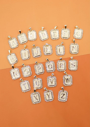 A complete set of gold rectangular alphabet letter charms from A to Z, featuring sparkling rhinestone borders, is artfully arranged on a warm peach background.