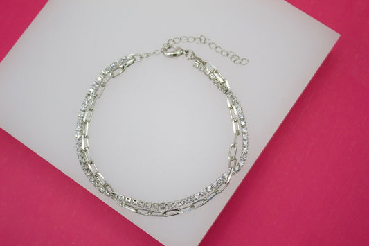 A double-strand silver tennis bracelet featuring interlocking link and crystal-encrusted chains rests on a white display surface against a vibrant pink background.