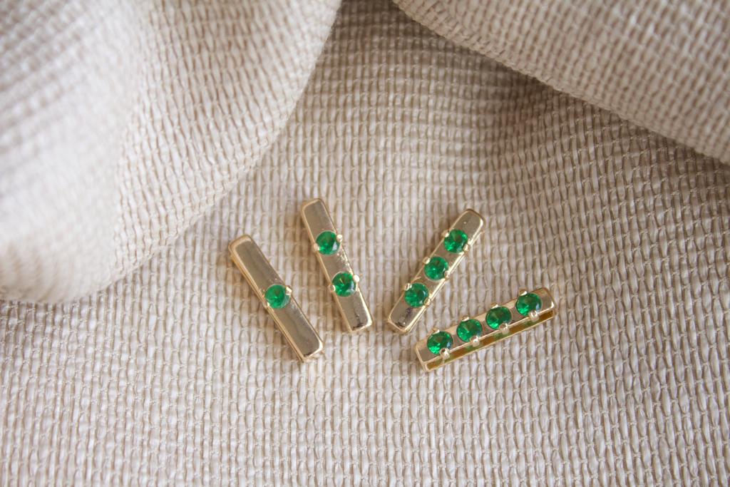 Four gold jewelry connectors set with brilliant green cubic zirconia stones rest on a textured beige fabric.