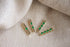 Four gold bar charms adorned with vibrant green gemstones rest on a textured beige fabric.