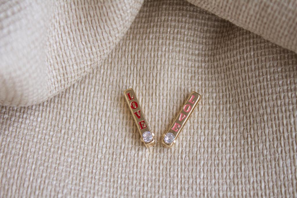 A pair of gold bar earrings featuring the word LOVE spelled out in red lettering and accented with sparkling cubic zirconia stones rests on a textured beige fabric.
