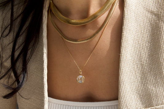 A stylish woman wears layered gold chain necklaces featuring a delicate pendant with a diamond-studded initial M.