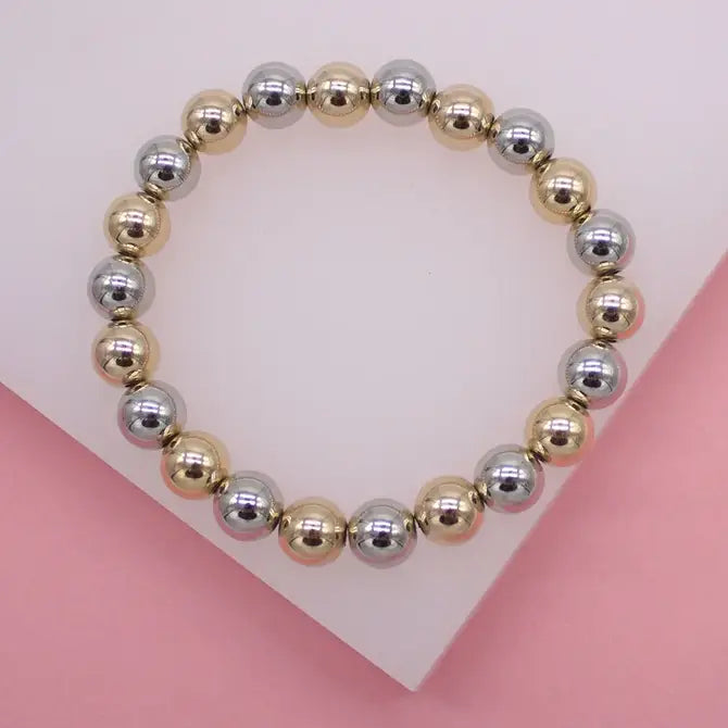 A beaded stretch bracelet features alternating metallic gold and silver spherical beads arranged in a circle on a white display stand.
