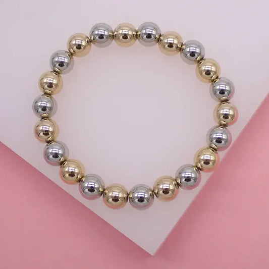 A beaded stretch bracelet features alternating metallic gold and silver spherical beads arranged in a circle on a white display stand.