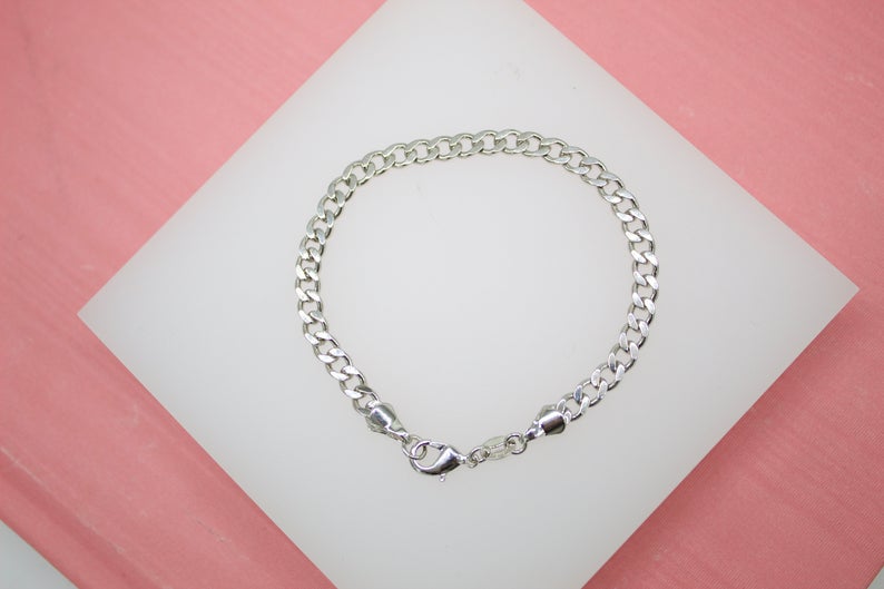 A polished silver curb chain bracelet lies displayed on a white stand against a pink background.