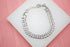 A silver cuban link chain bracelet rests on a white display stand against a pink background.