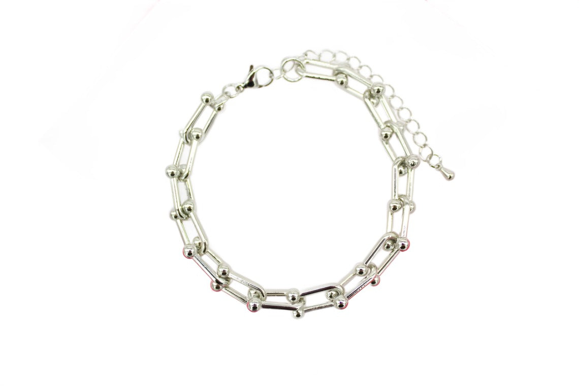 A shiny silver paperclip chain bracelet featuring interlocking links accented with small metallic beads lies curved against a clean white background.