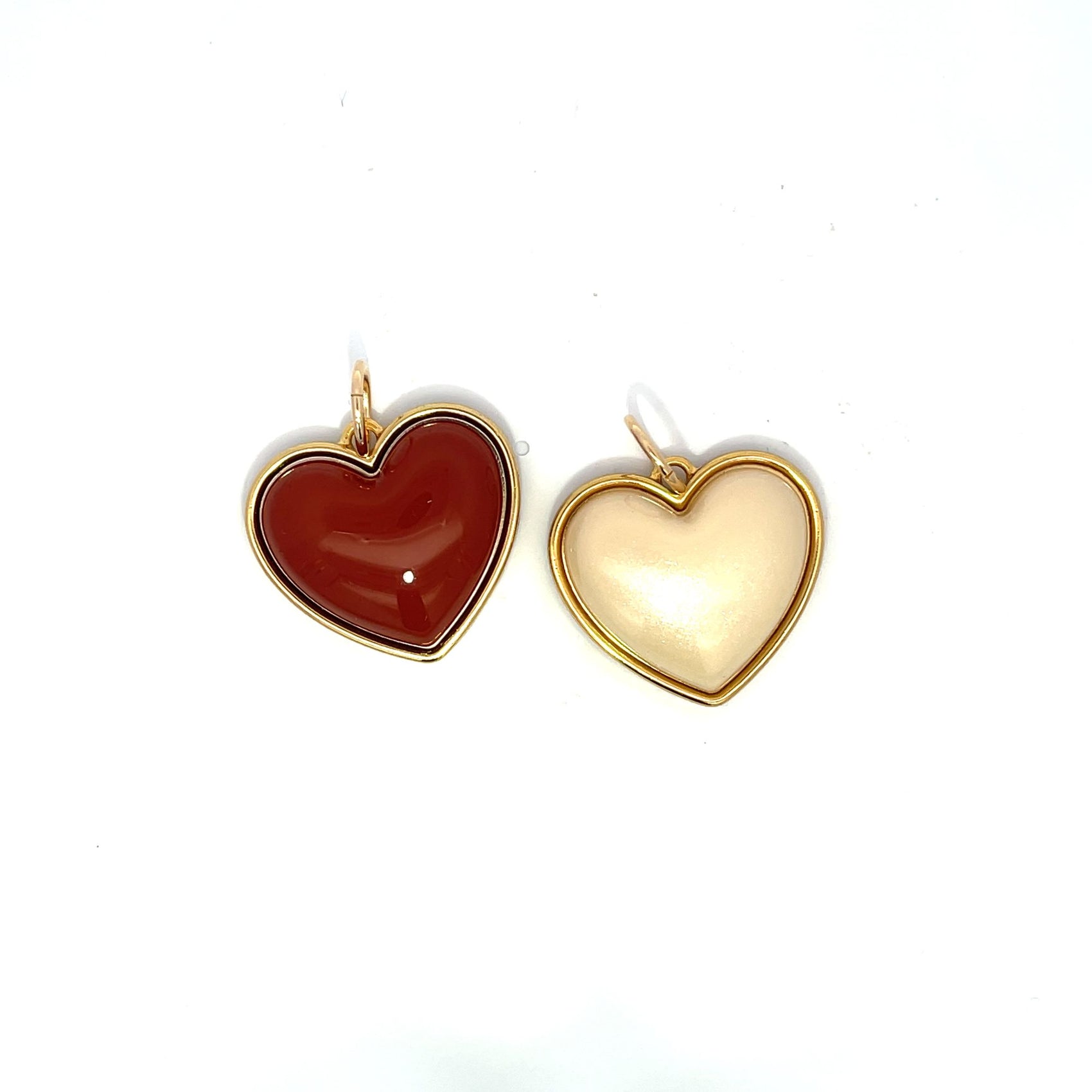 Two enamel heart-shaped pendants with gold borders rest against a white background, featuring one deep red charm and one creamy beige charm.