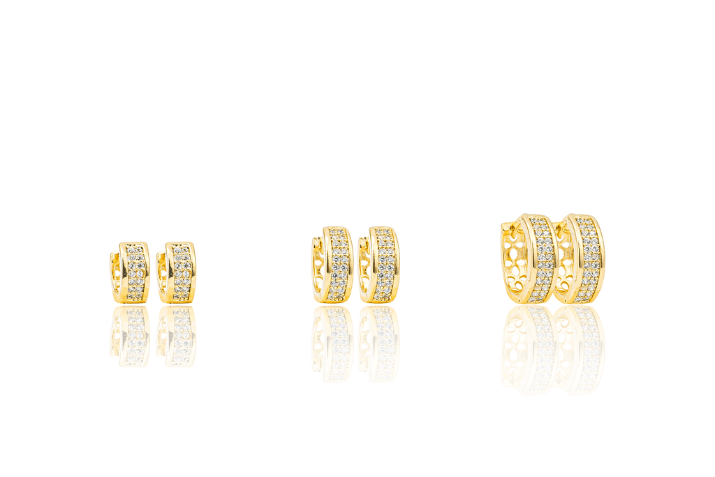 Three pairs of gold huggie hoop earrings encrusted with sparkling cubic zirconia stones display their reflections on a white surface.