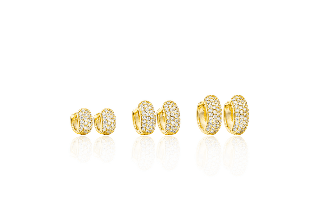 Three graduated pairs of pavé-set gold huggie hoop earrings sit neatly in a row, casting soft reflections on a white surface.