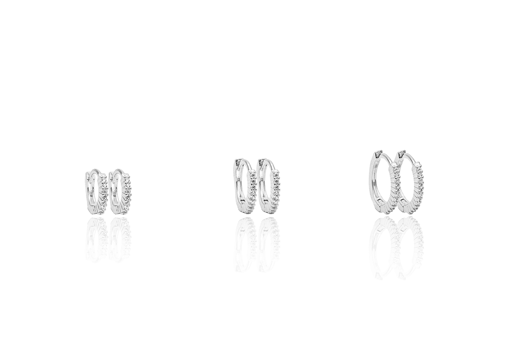Three pairs of sparkling silver huggie hoop earrings with CZ stones sit in graduated sizes on a reflective white surface.