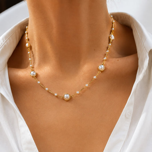 A woman wears a delicate gold chain necklace featuring alternating pearls and gold bar accents while dressed in a white button-down shirt.