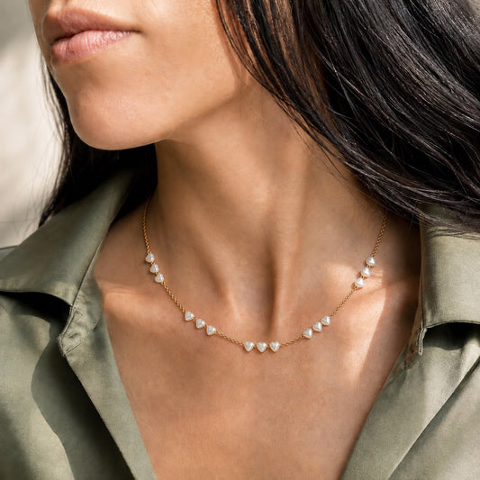 A woman wearing an olive-green button-down shirt showcases a delicate gold chain necklace featuring small pearl heart pendants.