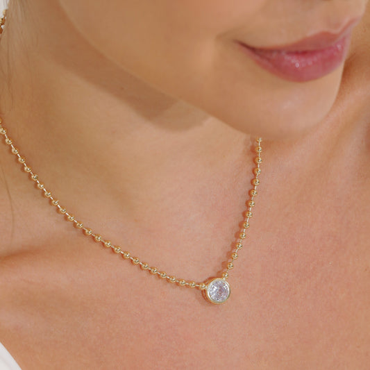 A woman wears a delicate gold beaded chain necklace featuring a sparkling bezel-set diamond pendant resting on her collarbone.