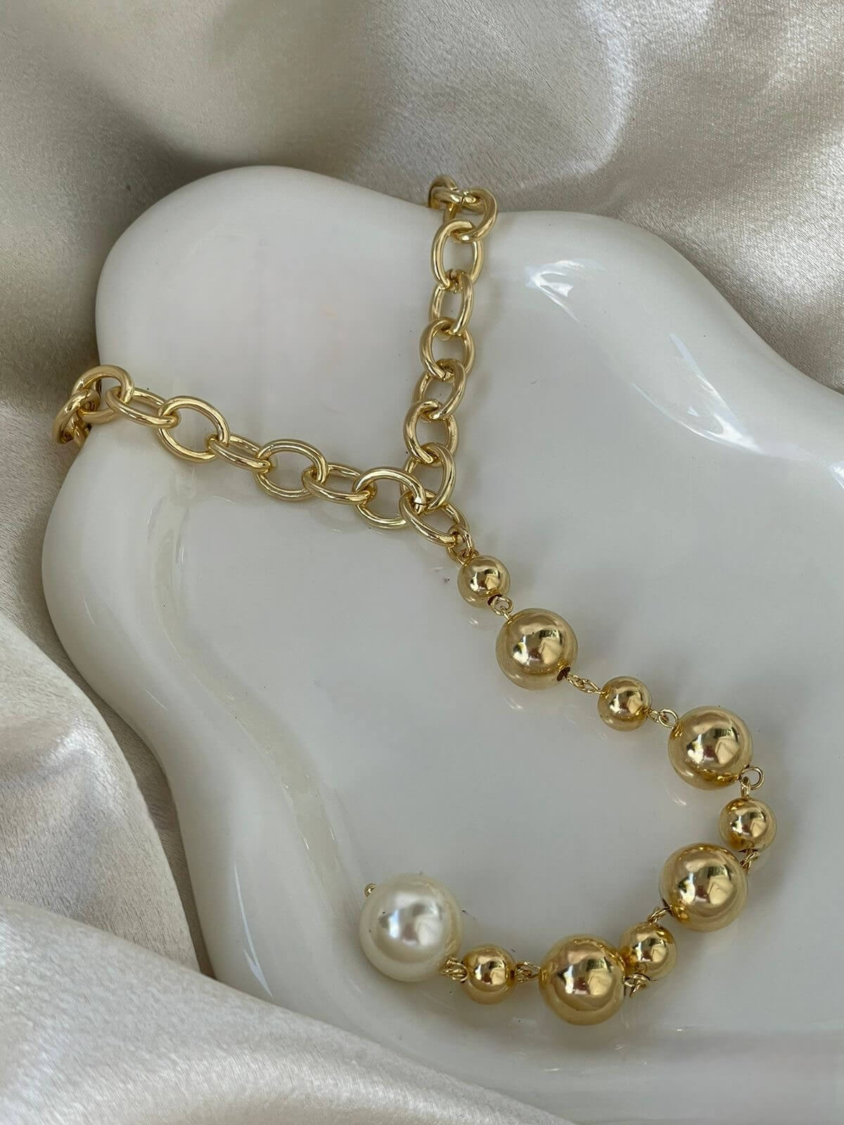 A chic gold chain necklace featuring assorted metallic beads and a single white pearl rests gracefully on a smooth white display surface.