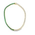 A gold tennis necklace features alternating emerald-cut green and white gemstones arranged in a continuous loop against a white background.