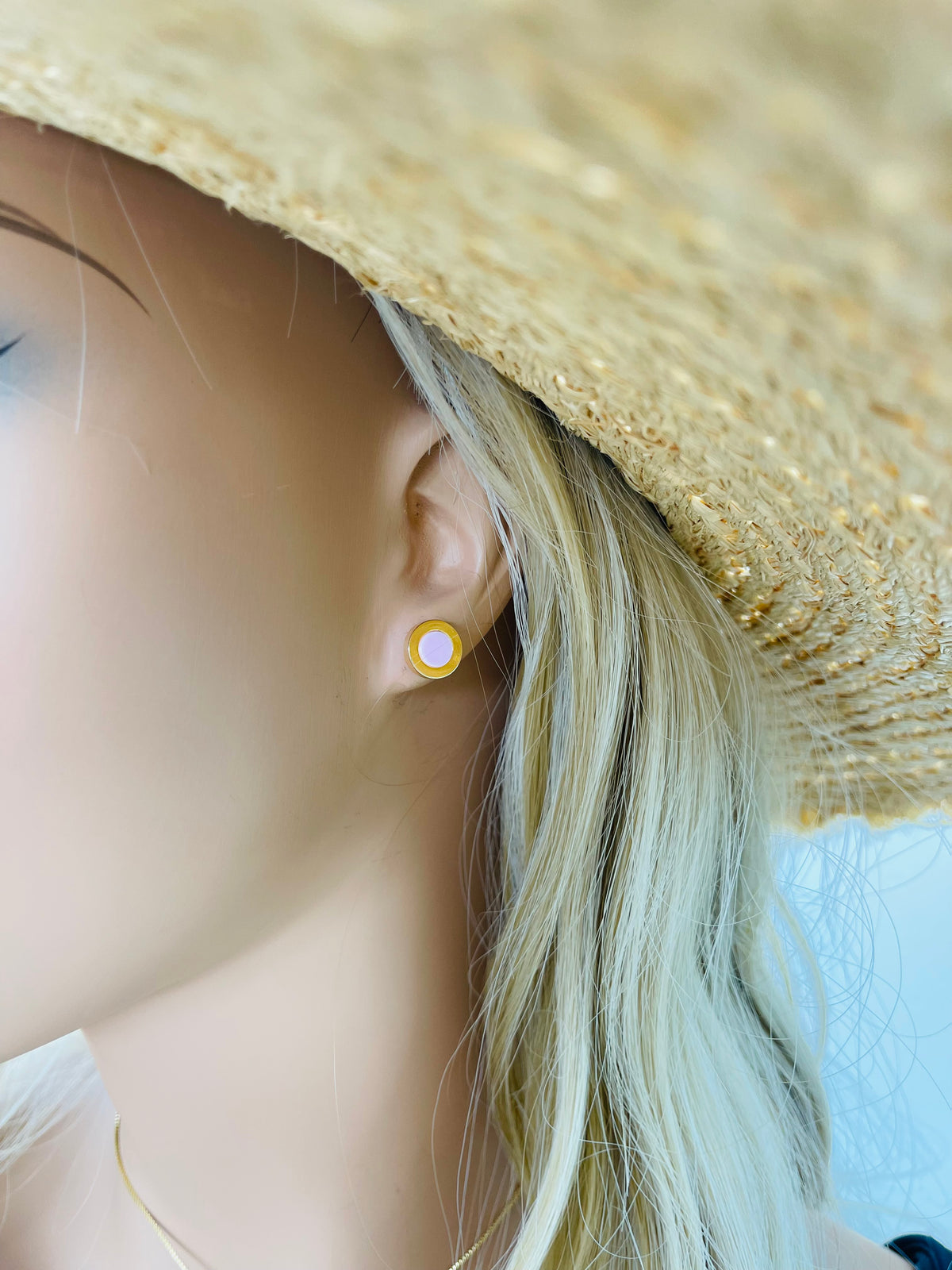 A mannequin wearing blonde hair and a straw sunhat displays a round gold enamel stud earring with a light pink center.