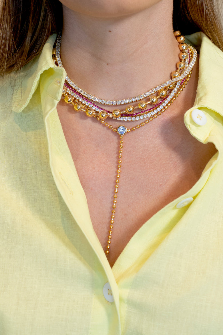 A close-up of a model wearing a multi-layered gold necklace with rhinestone and pink bead details, styled with a pale yellow button-down shirt.