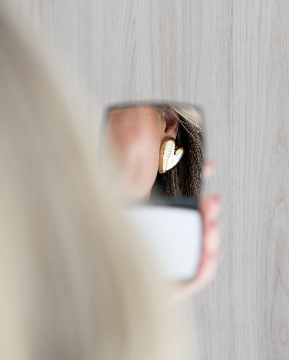 A woman holds a small mirror, reflecting her ear adorned with a chunky gold heart-shaped earring.