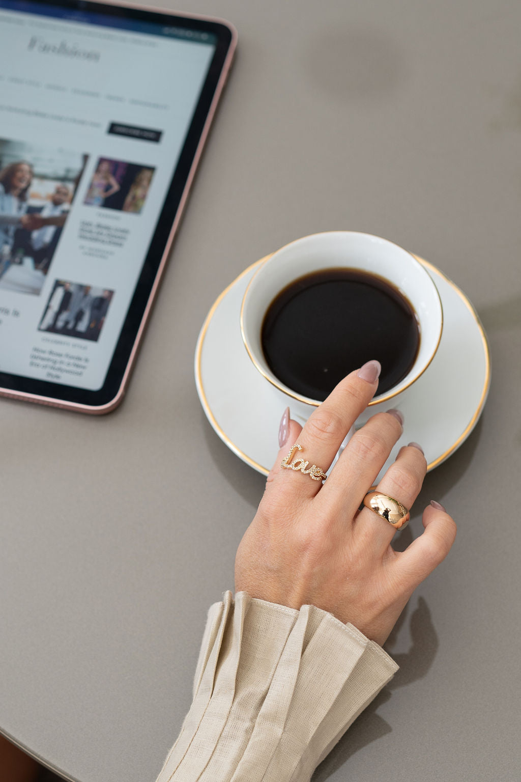 A woman wearing gold "Love" and dome rings reaches for a white coffee cup next to a tablet on a table.