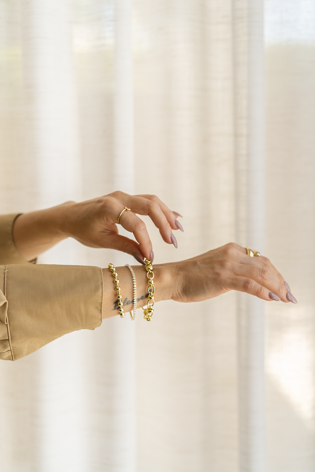 A woman wears a stack of delicate gold bracelets on her wrist, showcasing matching rings against a soft, neutral curtain backdrop.