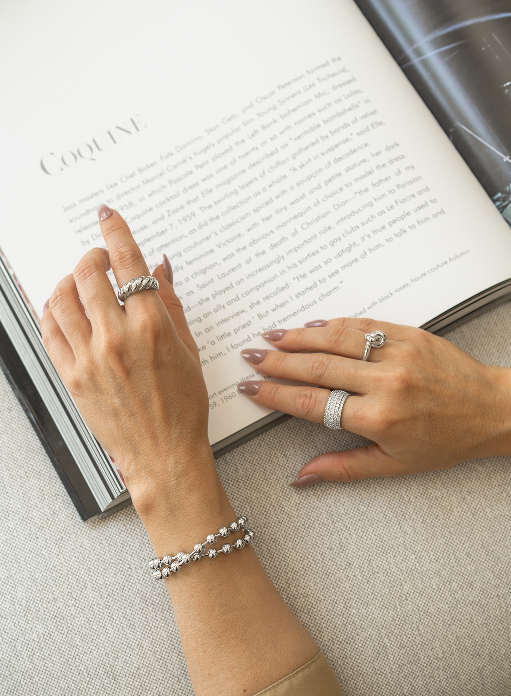 A woman with nude manicure showcases stylish silver jewelry, including a twisted rope ring, a knot ring, a pavé band, and a beaded double bracelet, while resting her hands over an open magazine.