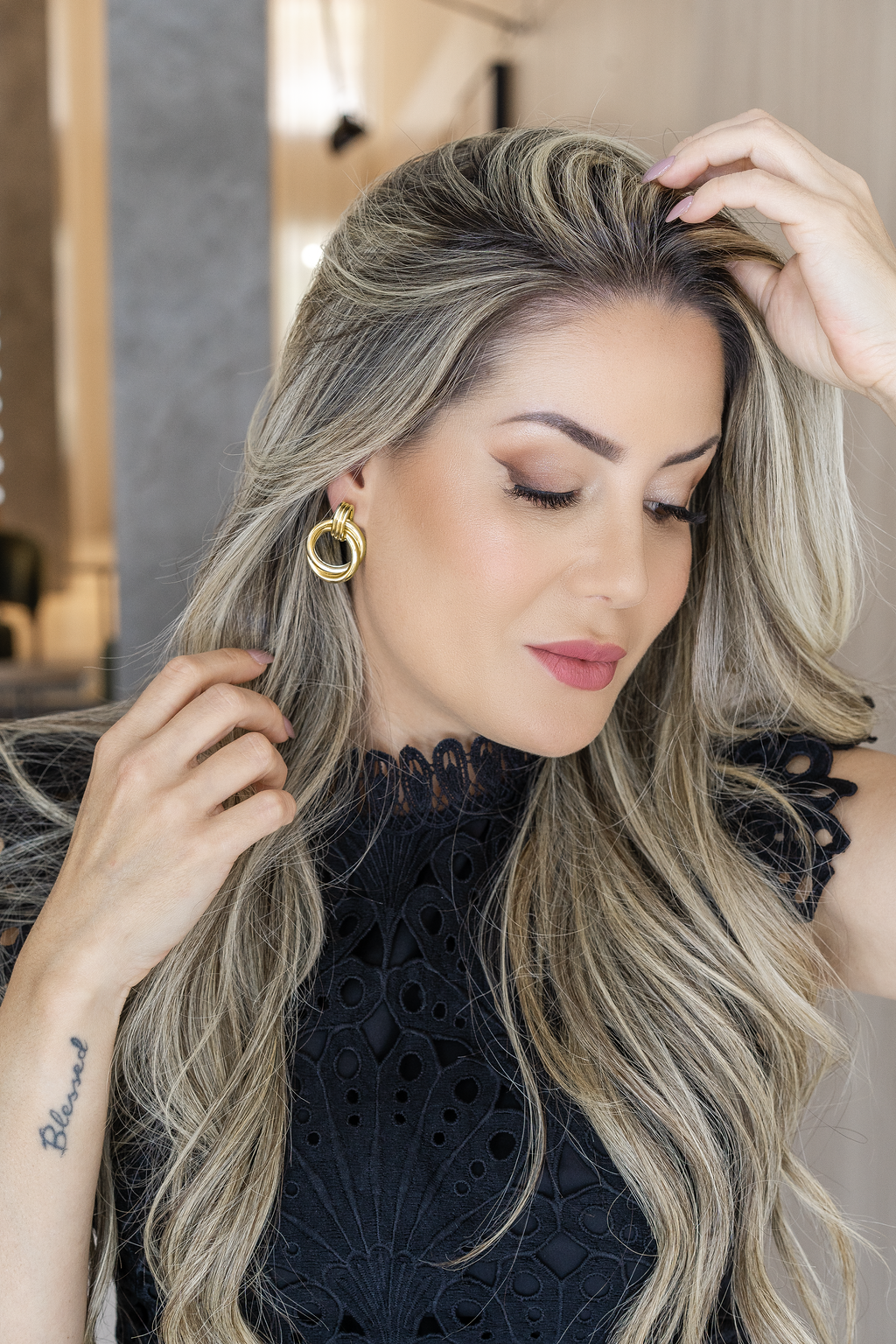 A blonde woman wearing a black lace top showcases a striking gold hoop earring while gently styling her highlighted hair.