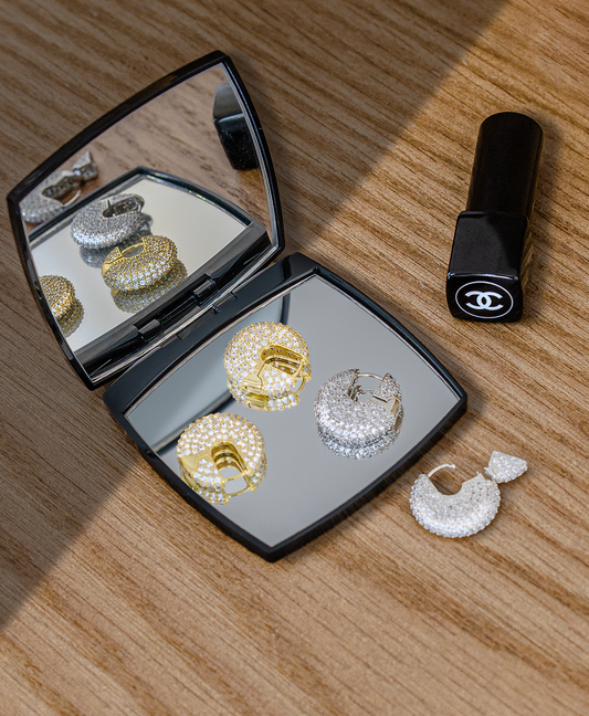 Pave-set gold and silver hoop earrings rest neatly inside and around an open compact mirror on a wooden surface.