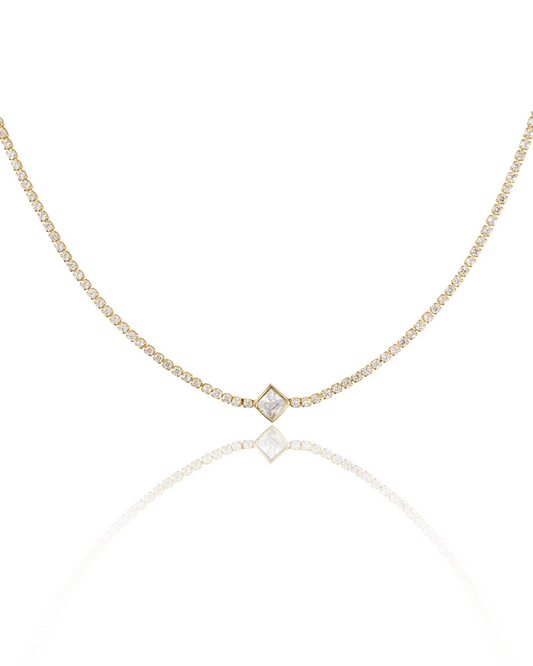 A delicate gold diamond tennis necklace featuring a square-cut pendant rests on a reflective white surface, displaying its sparkling stones.