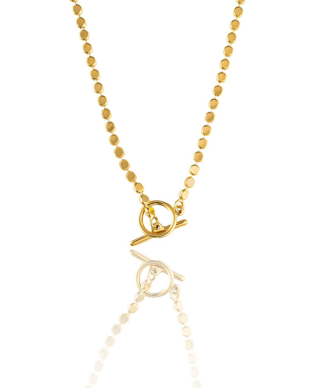 A delicate gold beaded necklace with a toggle clasp glimmers against a white reflective surface.