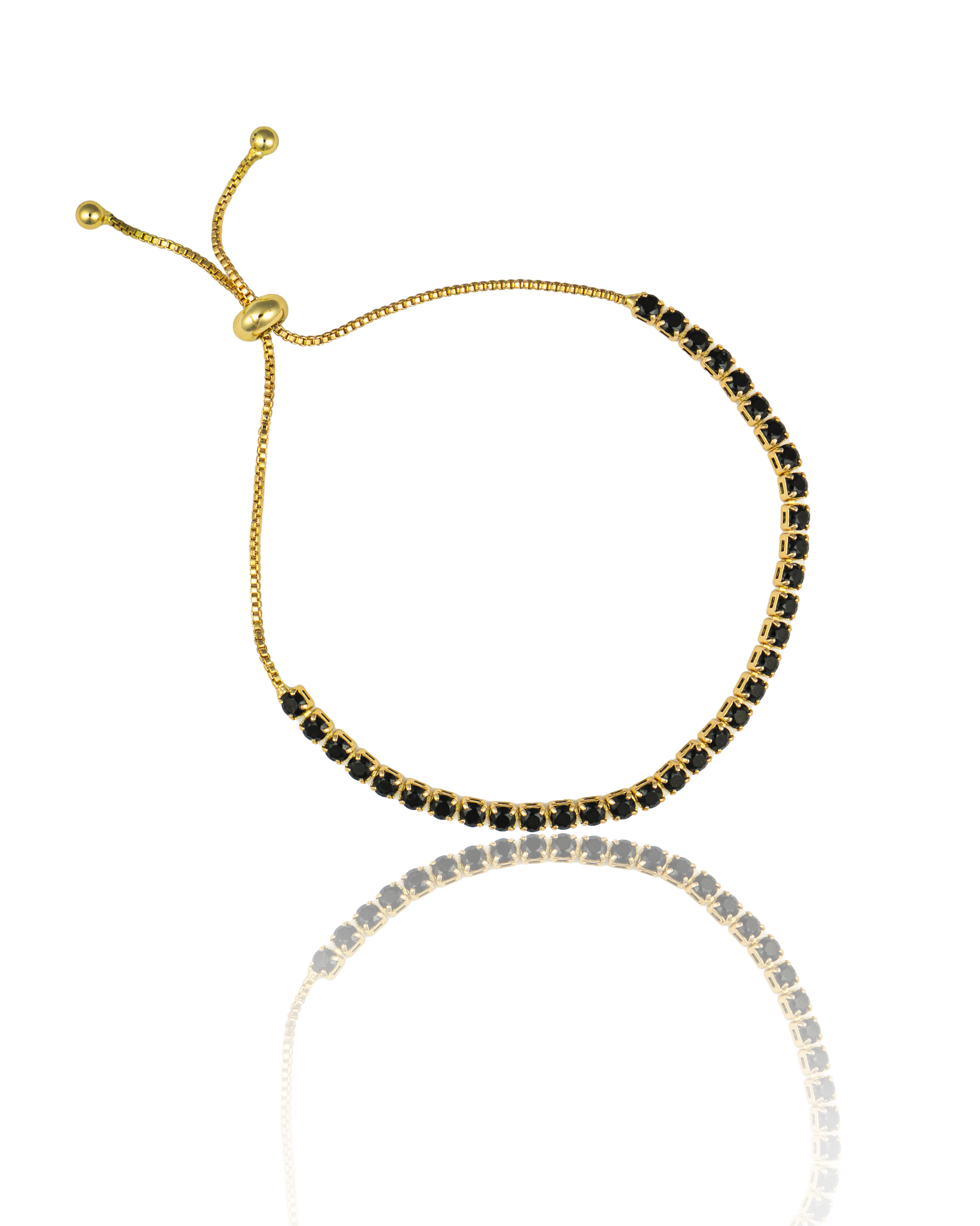 An elegant gold tennis bracelet featuring sparkling black cubic zirconia stones rests on a reflective white surface, showcasing its adjustable bolo clasp.