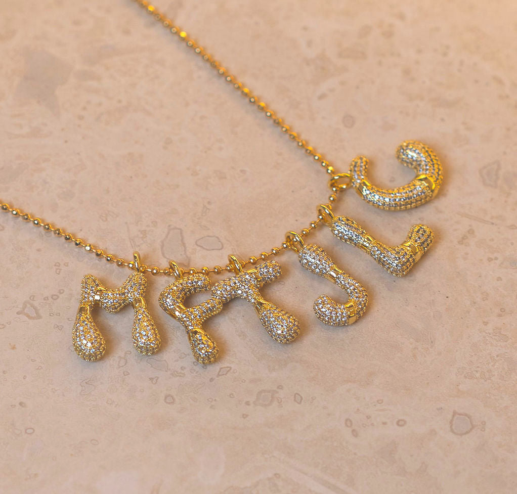 A gold-tone necklace with five dangling letter charms, each covered in small clear stones, displayed on a textured beige surface.