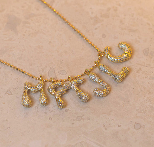 A gold-tone necklace with five dangling letter charms, each covered in small clear stones, displayed on a textured beige surface.