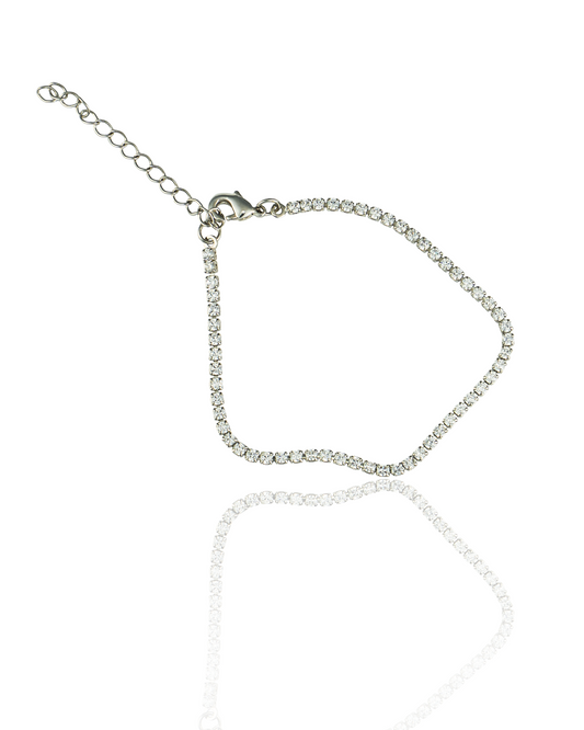 A sparkling silver rhinestone tennis bracelet lies on a reflective white surface, displaying its delicate crystal settings and adjustable chain clasp.