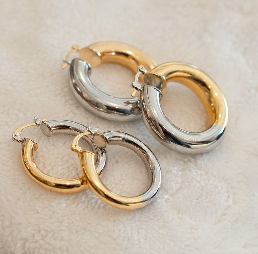 Two pairs of polished silver and gold hoop earrings arranged on a soft, textured off-white fabric background.
