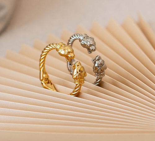 Three metallic cuff-style rings with sculpted animal head and body motifs, shown in gold and silver tones on a beige pleated paper background.