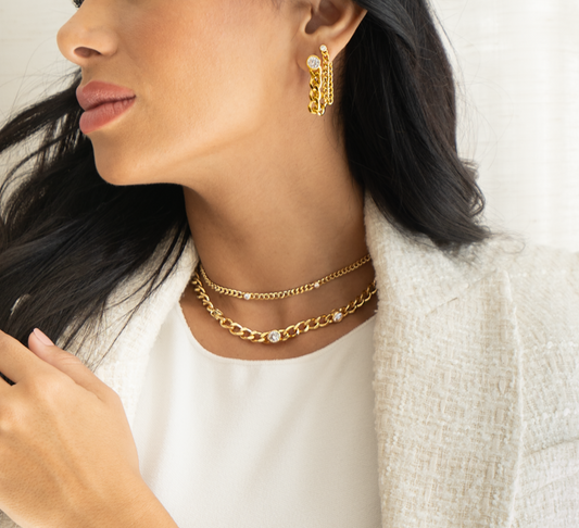 A stylish woman wears layered gold chain necklaces and matching statement earrings featuring cubic zirconia stones.