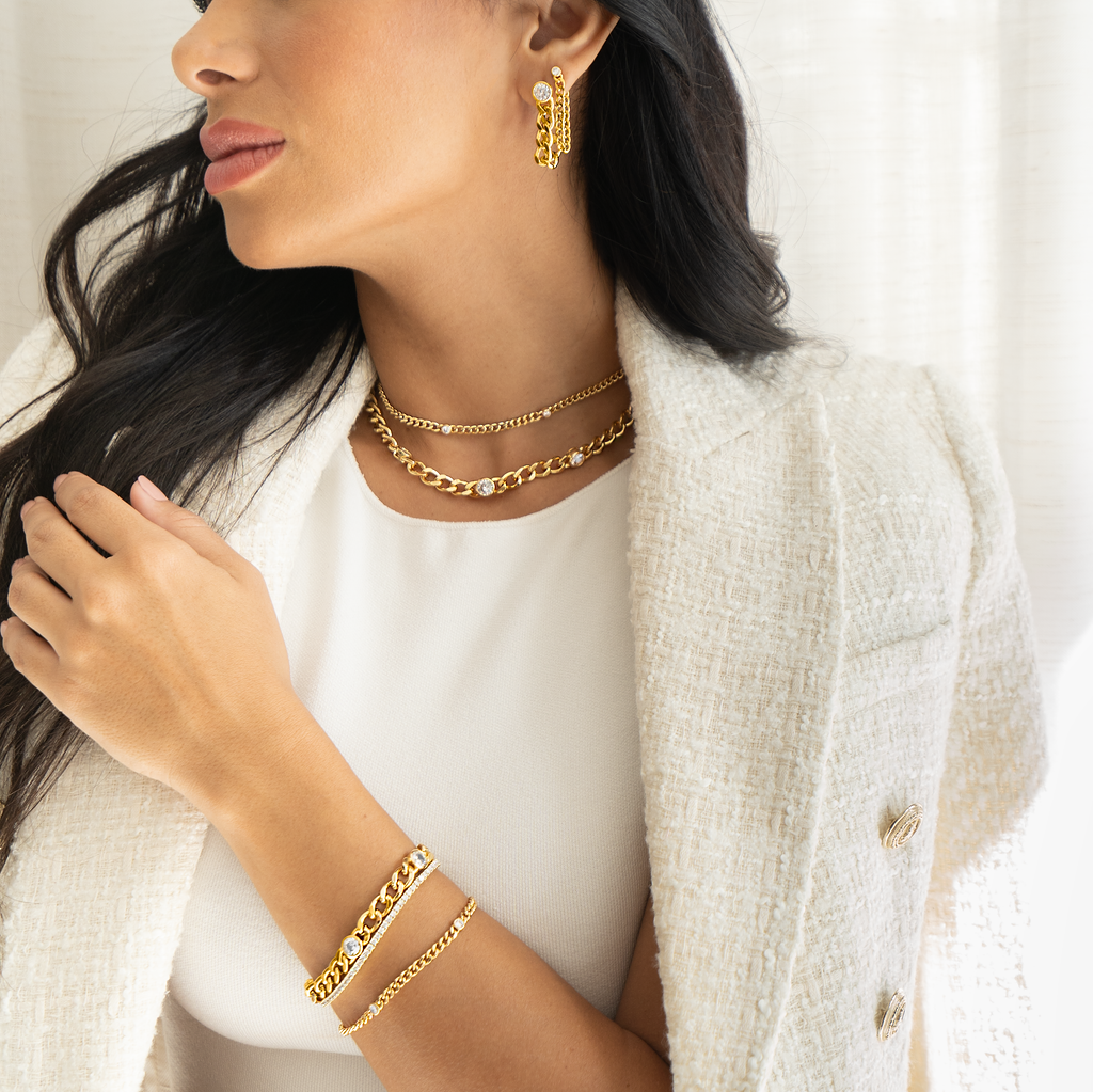 A stylish woman wearing a white tweed blazer showcases a matching set of gold chain jewelry, including layered necklaces, double-drop earrings, and stacked bracelets.