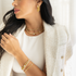 A stylish woman wearing a white tweed blazer showcases a matching set of gold chain jewelry, including layered necklaces, double-drop earrings, and stacked bracelets.