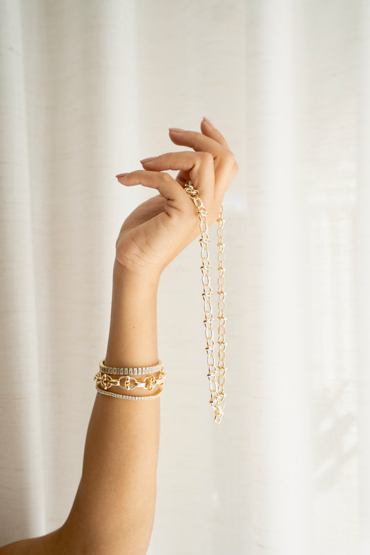 A woman holds up a chunky gold chain necklace while wearing matching gold link and diamond bracelets on her wrist against a light curtain background.