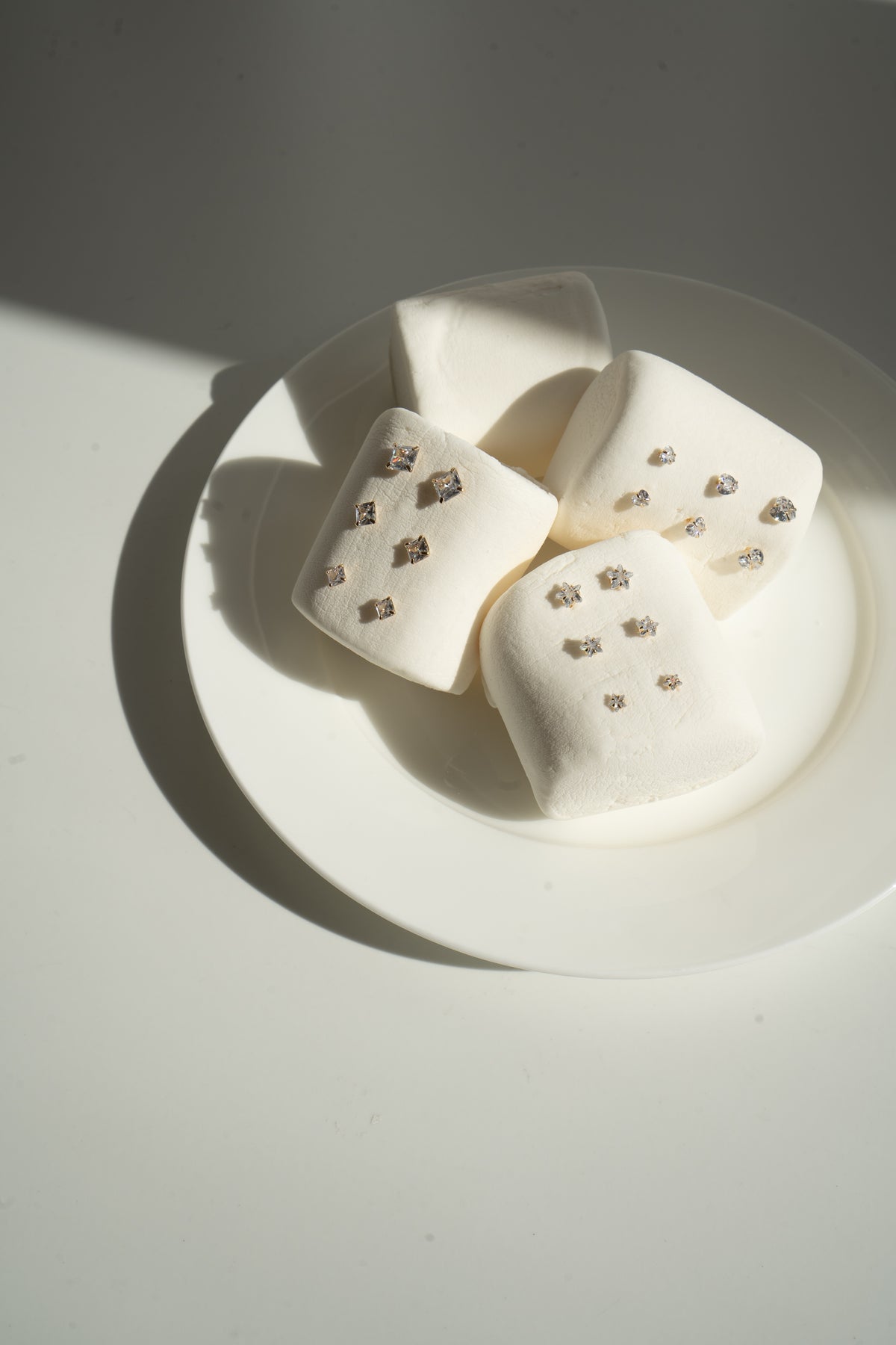 Four large white marshmallows rest on a white plate, artfully displaying sparkling gold stud earrings on their surfaces under soft sunlight.