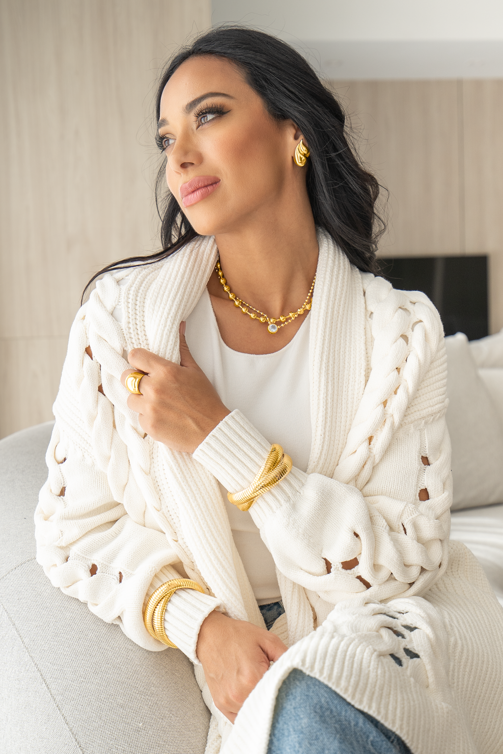 A stylish woman wearing an elegant gold jewelry set, including a statement necklace, earrings, rings, and stacked bracelets, sits indoors while modeling a cozy white knit cardigan.
