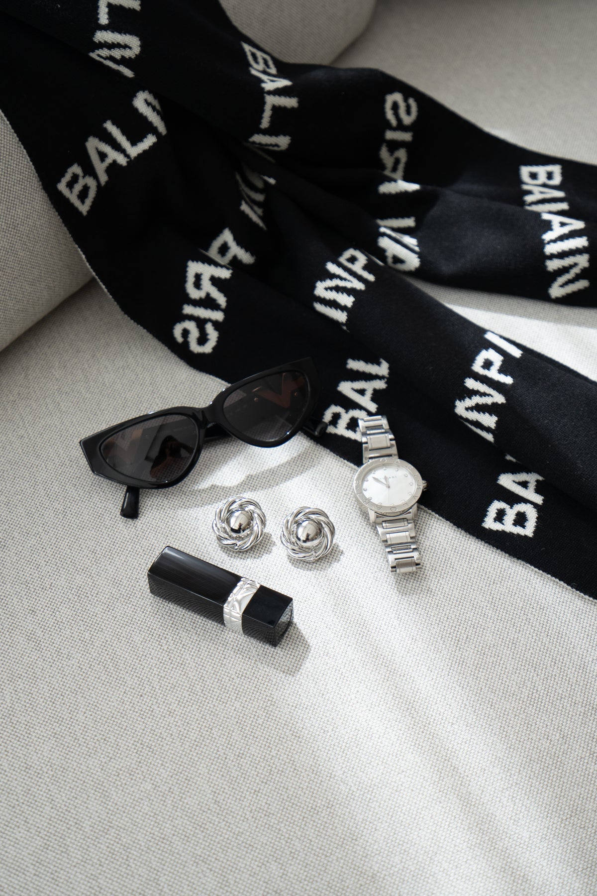 A stylish arrangement of luxury fashion accessories, including cat-eye sunglasses, a designer scarf, silver earrings, a wristwatch, and a lipstick tube, rests on a light-colored surface.