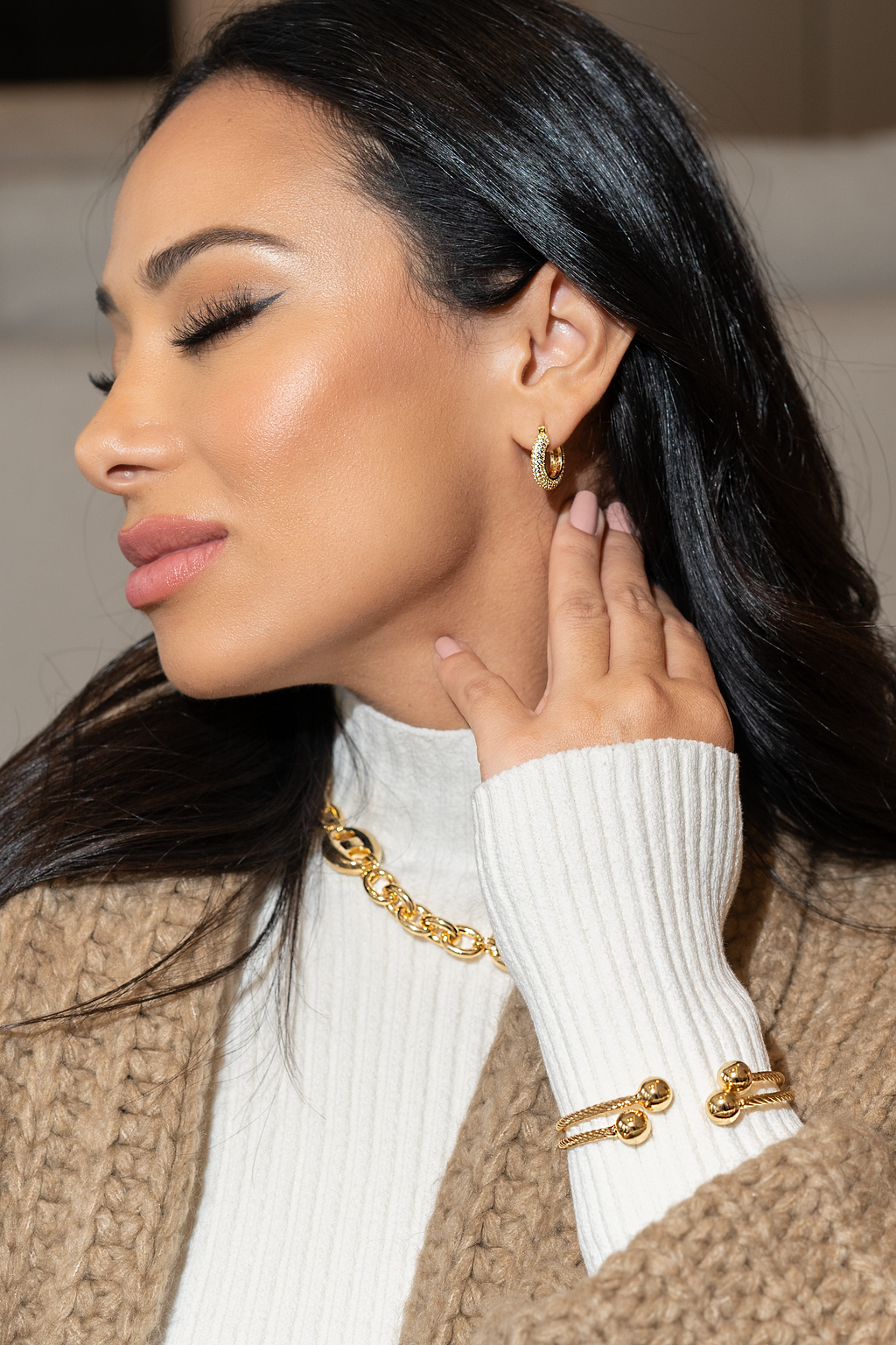 A glamorous woman wearing sparkling gold hoop earrings, a matching chain necklace, and a cable wire cuff bracelet gently touches her neck.