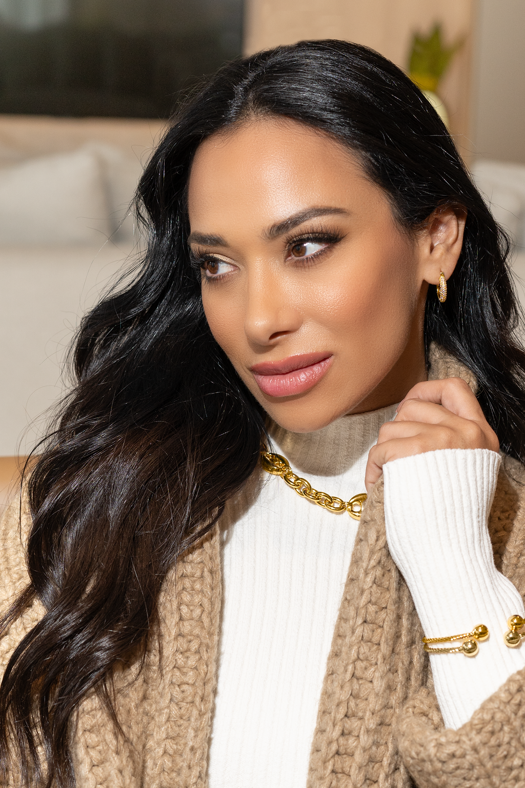 A stylish woman with dark, flowing hair wears a chunky gold chain necklace, gold hoop earrings, and a matching gold cuff bracelet while adjusting the sleeve of her cozy beige cardigan over a white turtleneck sweater.