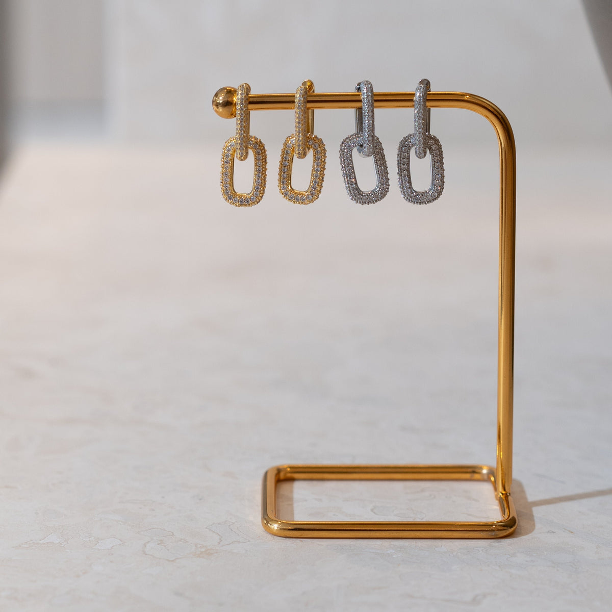 Four diamond-encrusted chain-link hoop earrings in gold and silver hang from a modern gold jewelry stand.