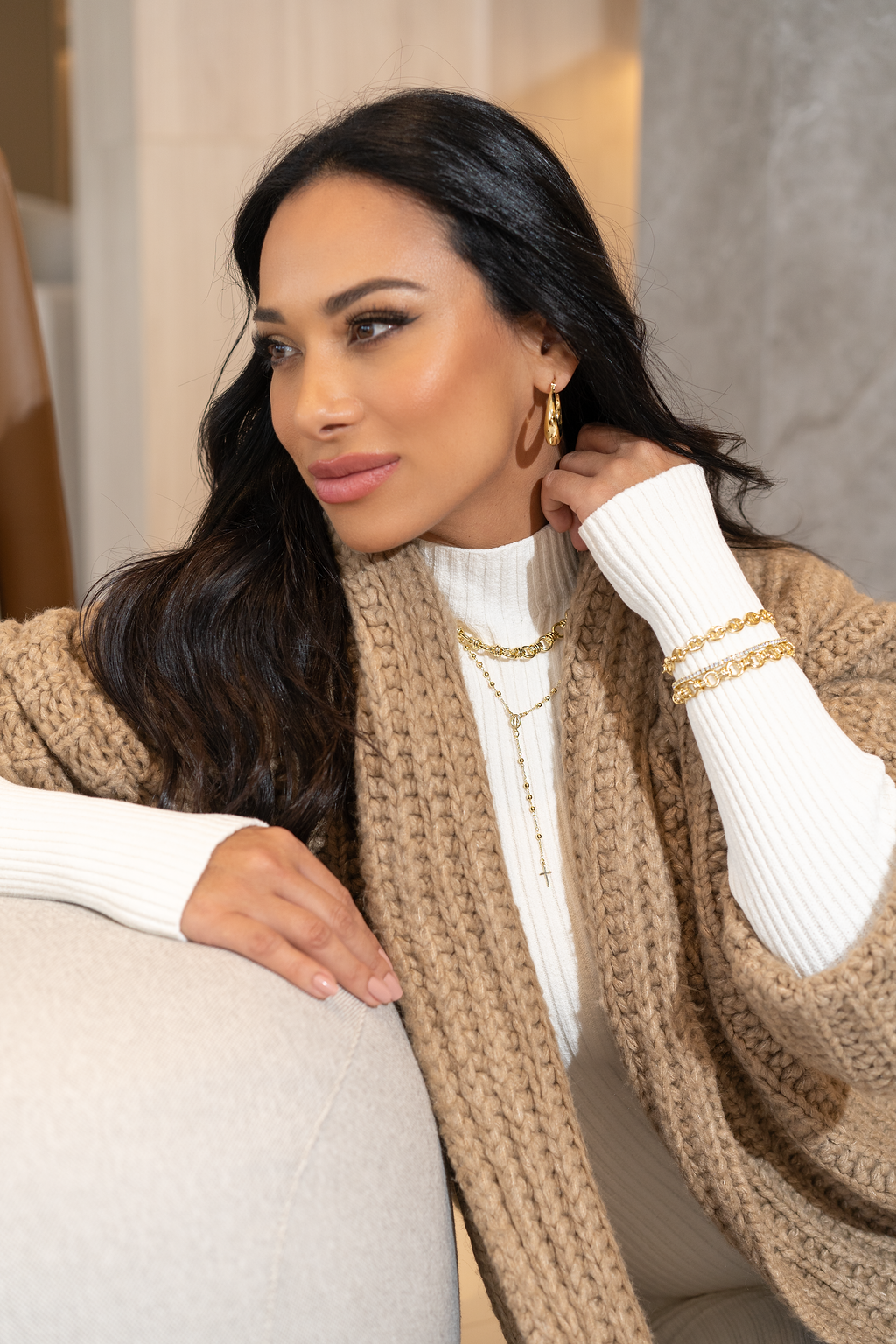 A stylish woman wearing gold layered necklaces, hoop earrings, and matching bracelets poses in a cozy tan cardigan and white turtleneck.