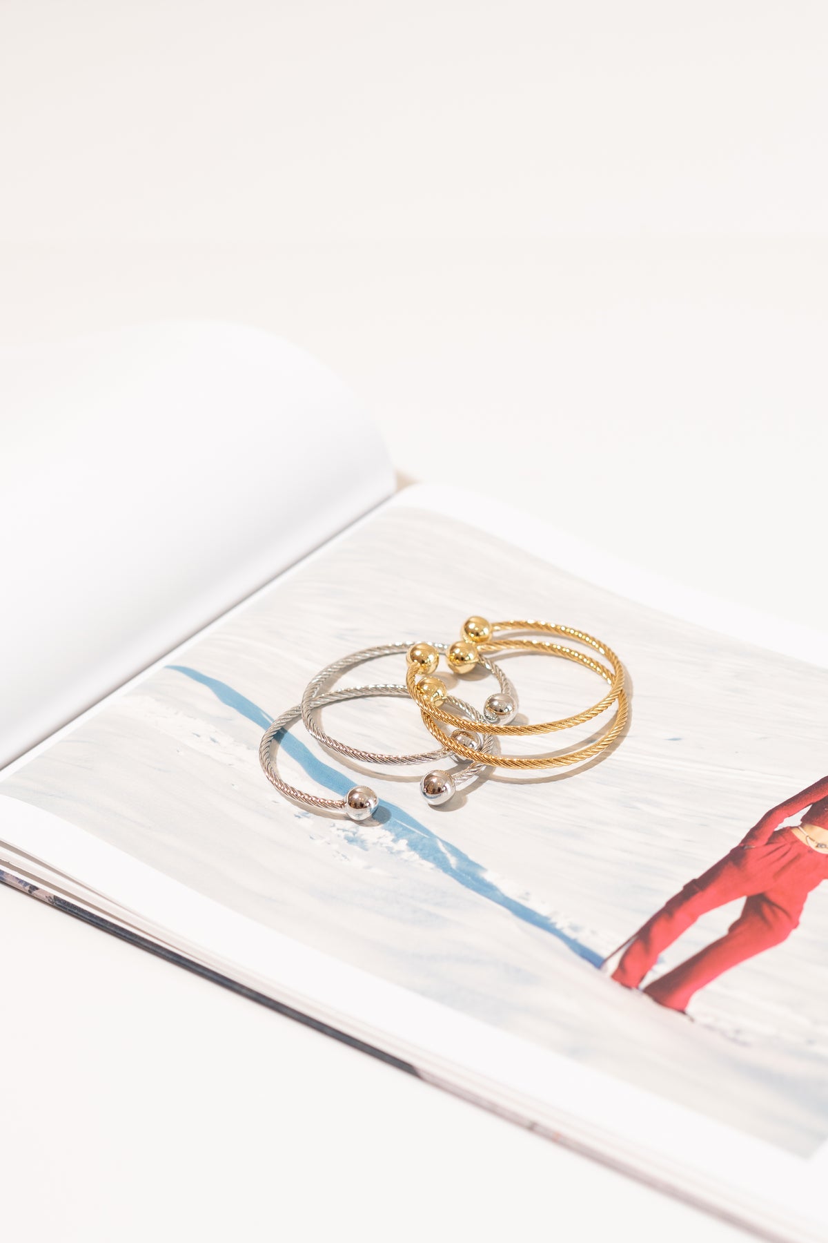 Two intertwined cable-wire bracelets featuring silver and gold tones rest on an open magazine page displaying a snowy landscape.