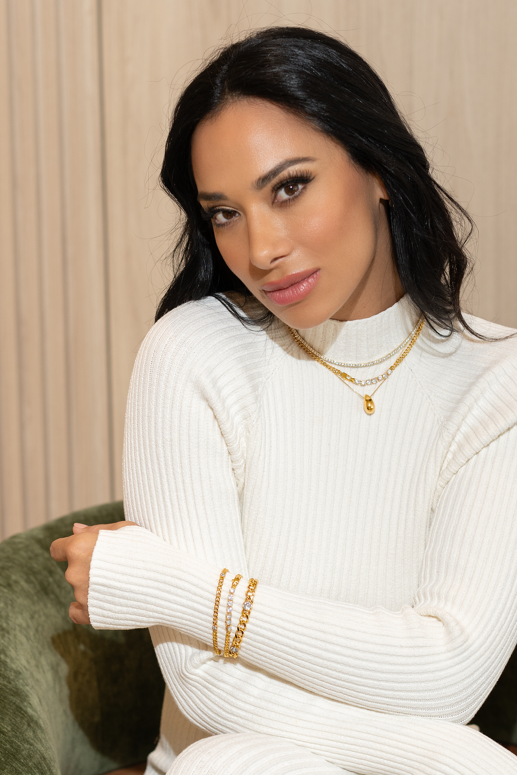 A stylish woman with dark hair wears a white ribbed sweater, layered gold necklaces, and gold bracelets while posing in a green armchair.