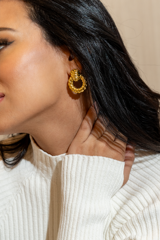 A woman with dark hair wears a textured gold hoop earring while gently touching her neck.